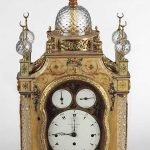 A large table clock with a cream and floral painted case, cut-glass columns, glass dome and spheres, with gilt-brass fittings including crescent moon finials