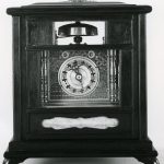 Small mantel clock; brass movement; pendulum; strike; revolving dial; double calendar; front-plate with geometric designs in colours; engraved front and back plates; turned baluster columns; rectangular base with musical box action; ebonised case with glazed sliding panel.